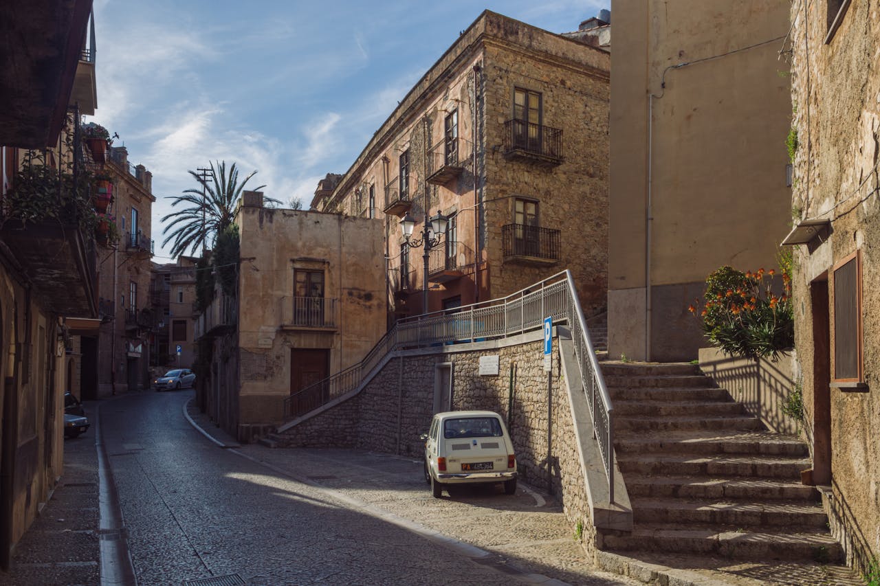 services-img Sunlit street view in Caccamo, Sicily with classic architecture and cars.